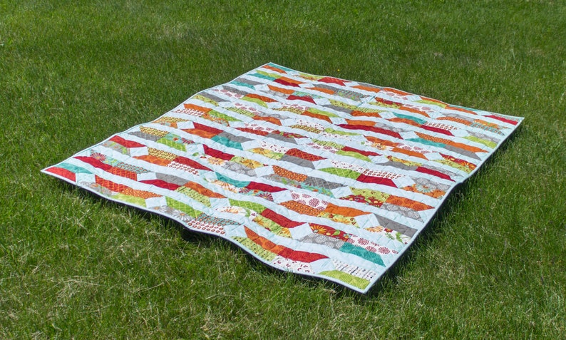 May include: A colorful patchwork quilt with a geometric pattern of arrows. The quilt is made with a variety of fabrics in shades of red, orange, yellow, green, blue, and white. The quilt is laid out on a green grassy surface.