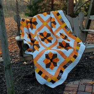 May include: A white, orange, and black quilt with a geometric pattern of flowers. The quilt is draped over a wooden swing set in a fall setting.