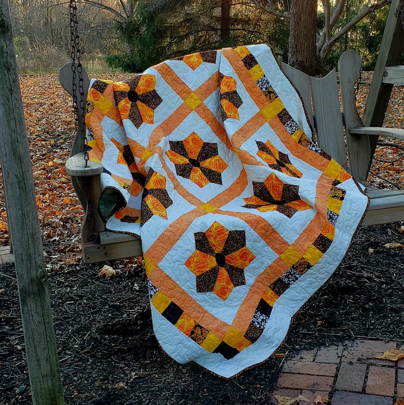 May include: A white, orange, and black quilt with a geometric pattern of flowers. The quilt is draped over a wooden swing set in a fall setting.