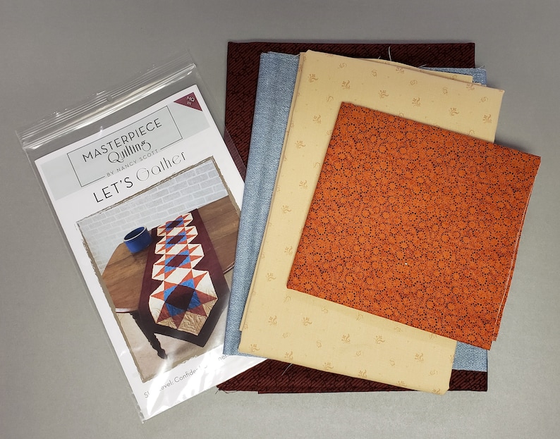 May include: A quilting kit for a table runner called "Let's Gather" by Nancy Scott. The kit includes fabric in shades of brown, orange, blue, and beige. The pattern is for a table runner with a geometric design.