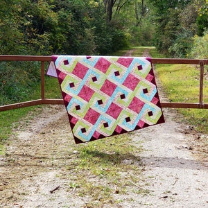 May include: A colorful quilt with a geometric pattern in shades of pink, green, and blue. The quilt is hanging on a wooden fence in a wooded area.