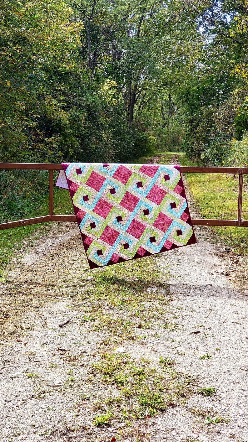 May include: A colorful quilt with a geometric pattern in shades of pink, green, and blue. The quilt is hanging on a wooden fence in a wooded area.