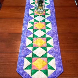 May include: A purple table runner with a geometric pattern of green, pink, and yellow stars. The runner is on a wooden table with a vase of flowers in the center.