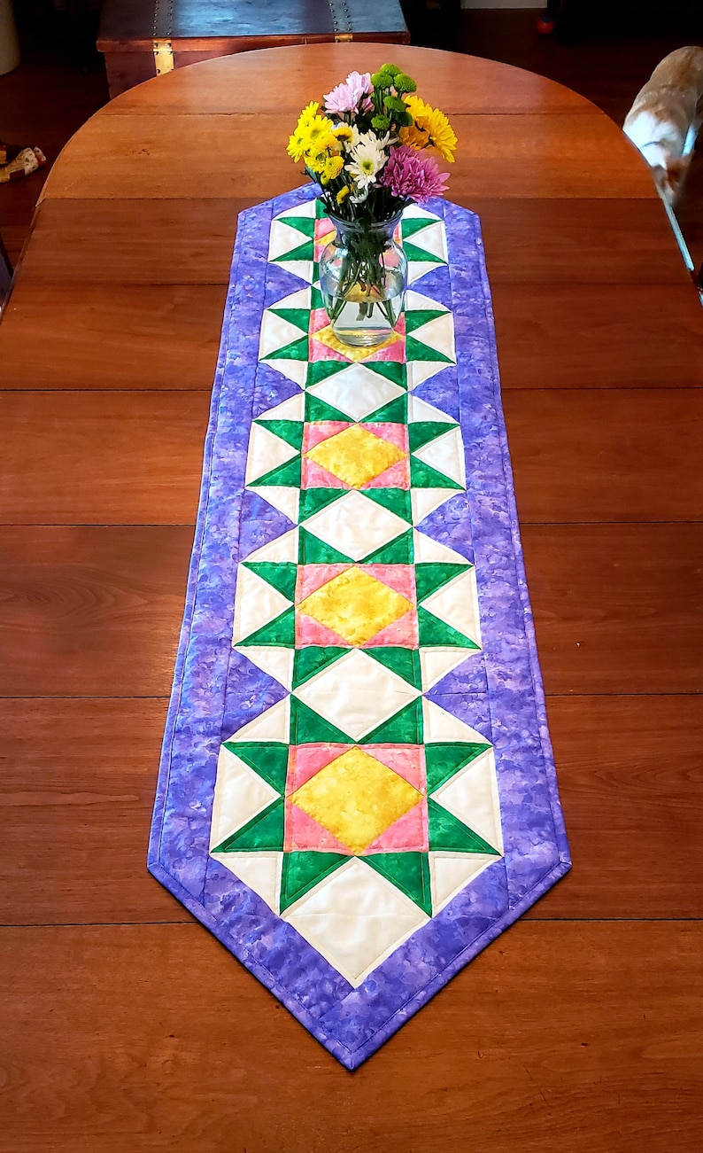 May include: A purple table runner with a geometric pattern of green, pink, and yellow stars. The runner is on a wooden table with a vase of flowers in the center.