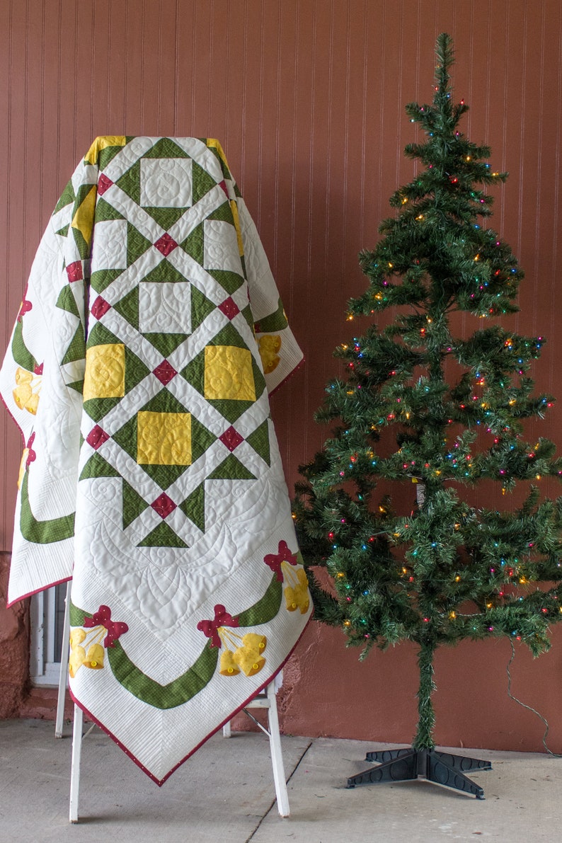 May include: A white, green, red, and yellow quilt with a bell pattern is draped over a white folding ladder. A decorated Christmas tree with colorful lights is in the background.