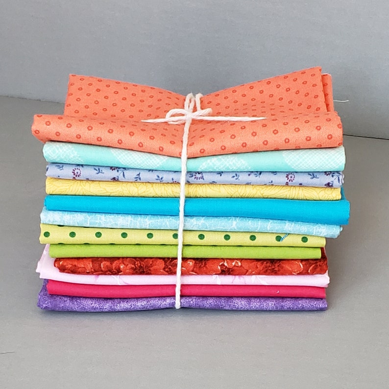 May include: A stack of 10 fabric squares in various colors and patterns, tied together with a white string. The fabrics include orange with white polka dots, light blue with white floral print, yellow with green floral print, turquoise, green with white polka dots, red with white floral print, pink with white floral print, and purple.