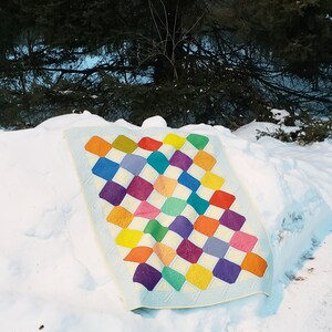 May include: A colorful quilt with a diamond pattern in shades of yellow, orange, red, purple, blue, and green. The quilt is made of fabric squares sewn together and has a light blue background.