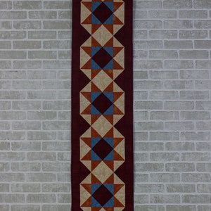 May include: A burgundy table runner with a geometric pattern of stars, diamonds, and squares in shades of orange, brown, blue, and cream.