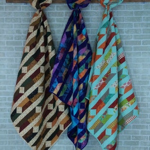 May include: Three handmade quilts hanging on a brick wall. The quilt on the left is brown and tan with a geometric pattern. The quilt in the middle is purple and blue with a geometric pattern. The quilt on the right is turquoise and orange with a geometric pattern.