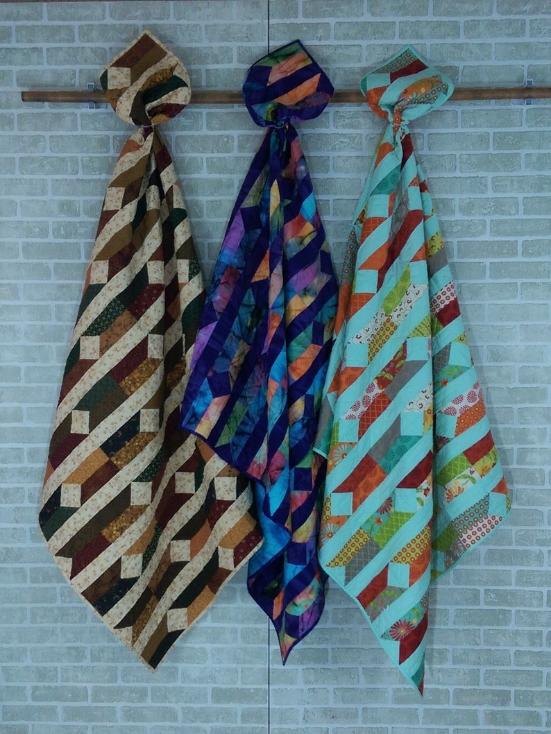 May include: Three handmade quilts hanging on a brick wall. The quilt on the left is brown and tan with a geometric pattern. The quilt in the middle is purple and blue with a geometric pattern. The quilt on the right is turquoise and orange with a geometric pattern.