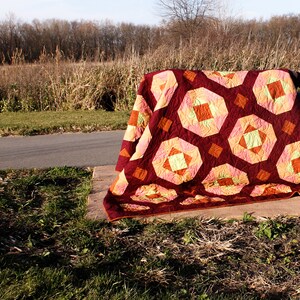May include: A colourful quilt with a geometric pattern in shades of red, orange, yellow, and green is draped over a wooden bench. The quilt is made of fabric squares sewn together to create a patchwork design.