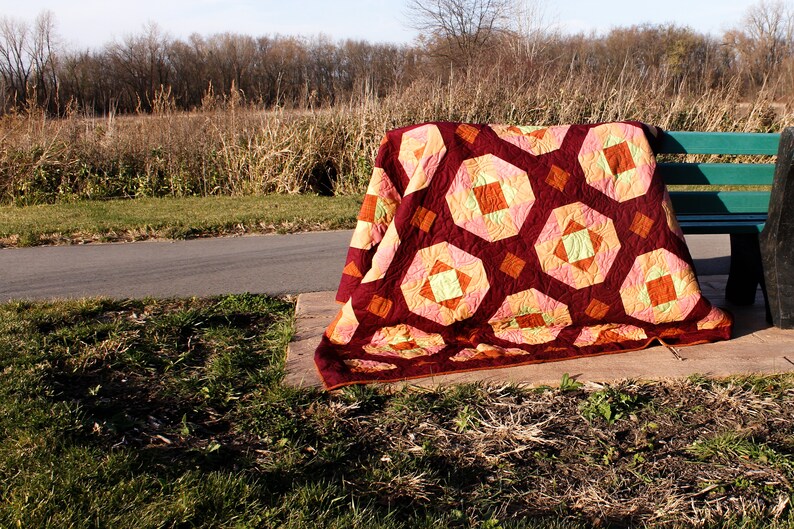 May include: A colourful quilt with a geometric pattern in shades of red, orange, yellow, and green is draped over a wooden bench. The quilt is made of fabric squares sewn together to create a patchwork design.