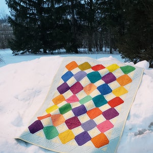 May include: A colorful quilt with a geometric pattern of squares and diamonds. The quilt is made with a variety of bright colors, including red, orange, yellow, green, blue, purple, and pink. The quilt is lying on a bed of snow.