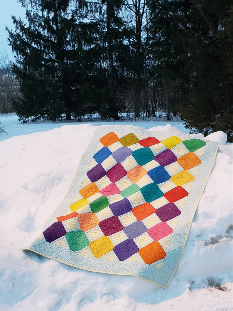 May include: A colorful quilt with a geometric pattern of squares and diamonds. The quilt is made with a variety of bright colors, including red, orange, yellow, green, blue, purple, and pink. The quilt is lying on a bed of snow.