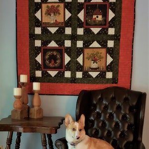 May include: A white and orange corgi dog sitting in a brown leather armchair in front of a wall with a red, green, and white quilt hanging on it. There are three wooden candle holders on a wooden table in front of the chair.