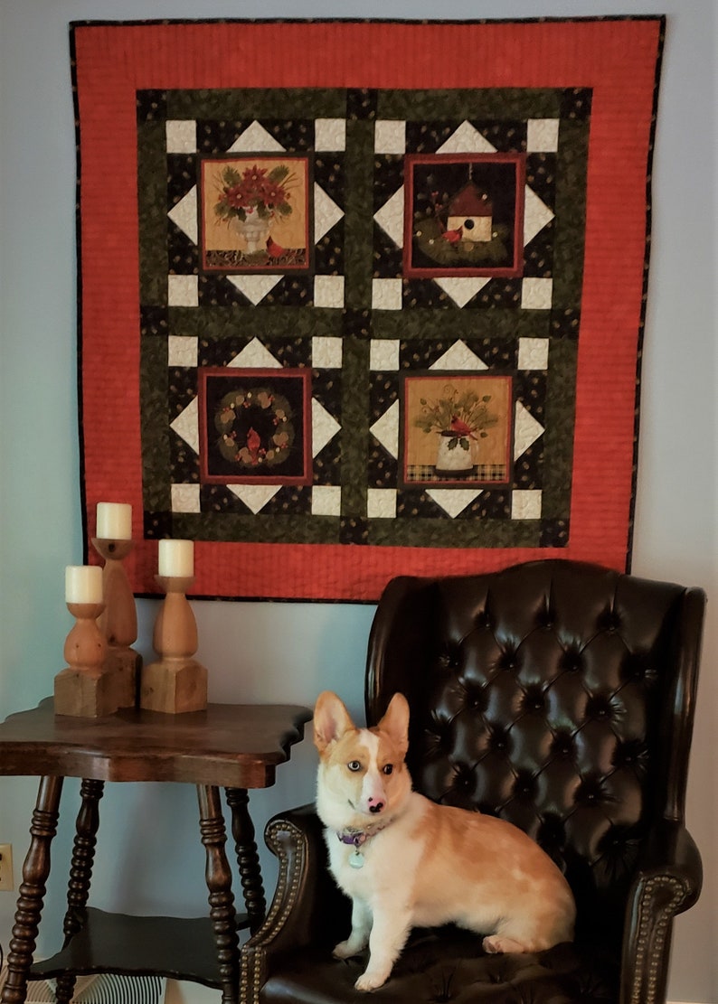 May include: A white and orange corgi dog sitting in a brown leather armchair in front of a wall with a red, green, and white quilt hanging on it. There are three wooden candle holders on a wooden table in front of the chair.