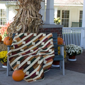 May include: A brown, green, and cream patchwork quilt is draped over a wooden bench with a fall theme. The bench is on a porch with pumpkins, hay bales, and mums.
