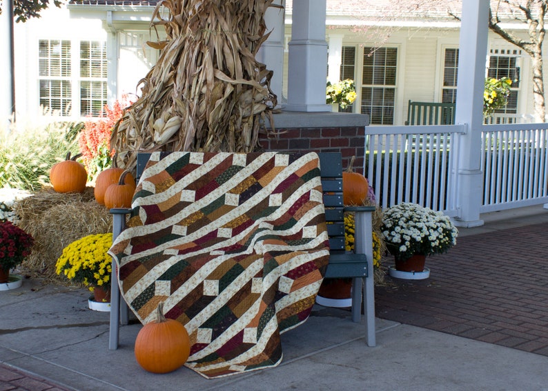 May include: A brown, green, and cream patchwork quilt is draped over a wooden bench with a fall theme. The bench is on a porch with pumpkins, hay bales, and mums.