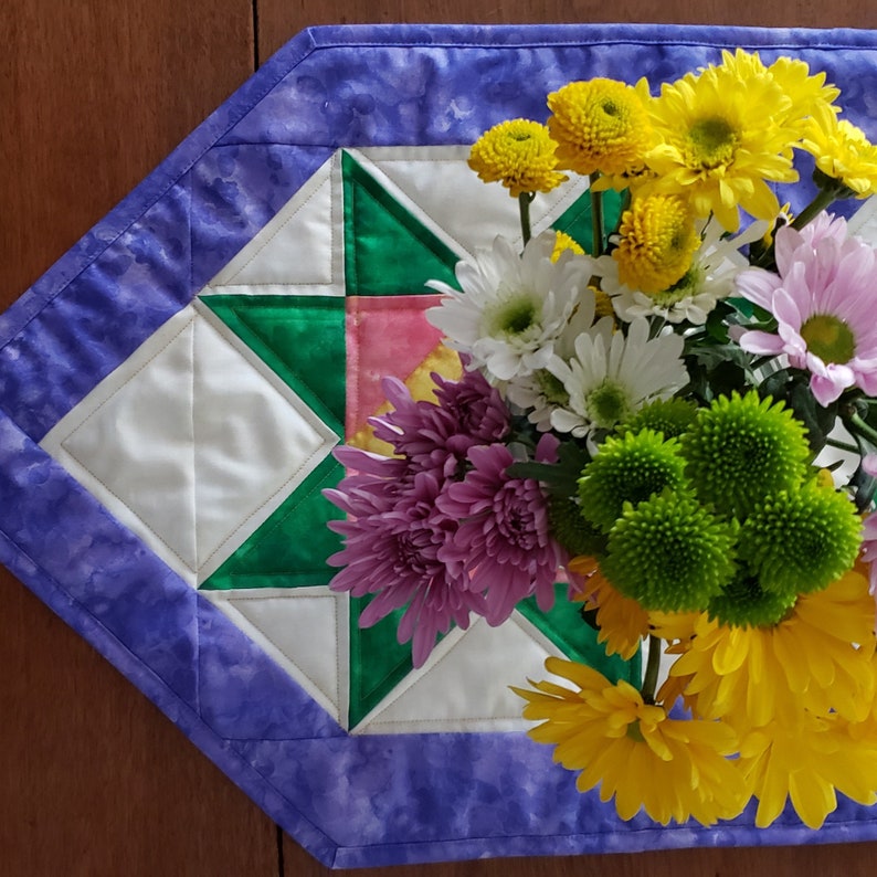 May include: A purple and white quilted table runner with a star design in the center. A bouquet of yellow, white, and purple flowers sits on top of the runner.