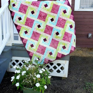 May include: A quilt with a geometric pattern in shades of pink, green, and blue. The quilt is hanging on a white railing on a porch.