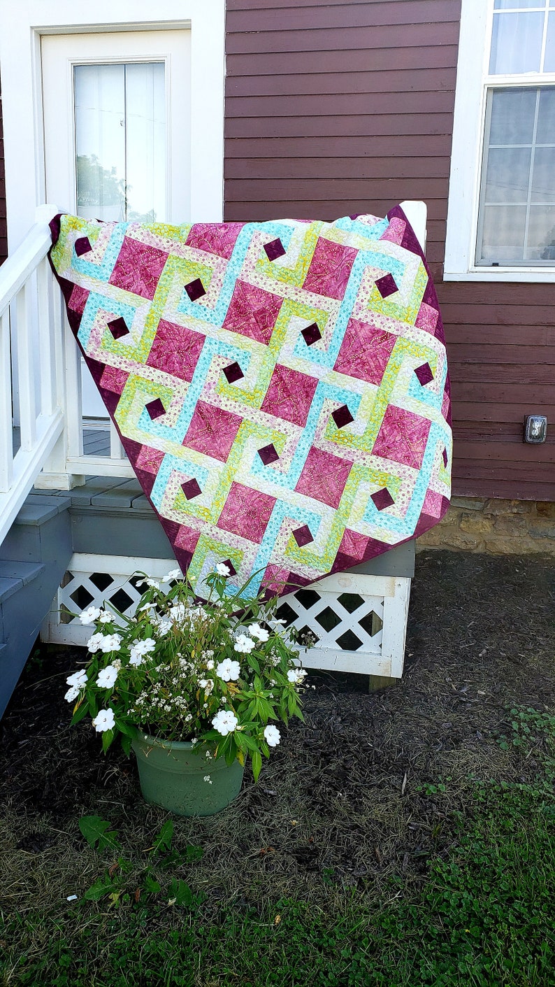 May include: A quilt with a geometric pattern in shades of pink, green, and blue. The quilt is hanging on a white railing on a porch.