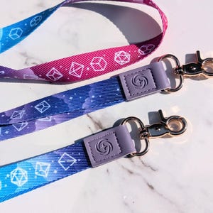 May include: Two lanyards with a dice pattern in shades of blue, purple, and pink. Each lanyard has a lavender-colored leatherette tab with a silver-colored clasp. The lanyards are designed for holding keys or ID badges.