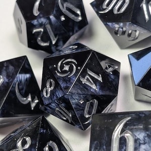 May include: A set of seven polyhedral dice for tabletop games. The dice are black with a marbled, smoky effect and silver numbering. The dice are arranged on a white surface.