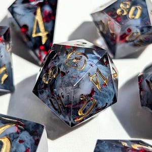 Può includere: Un set di sette dadi poliedrici caratterizzati da una base in resina marmorizzata bianca e nera con scaglie di glitter rosso incorporate e numeri incisi color oro. La collezione include varie forme geometriche per giochi da tavolo e di ruolo.