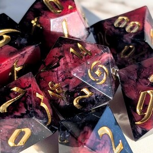 May include: A collection of polyhedral dice with a marbled red and black design. The dice have gold-colored numbers and are made of a clear resin material. The dice are for tabletop games.