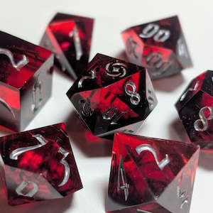 Pode incluir: Sete dados poliédricos com uma aparência vermelha profunda, marmoreada e numeração prateada. Os dados são projetados para jogos de RPG de mesa. Os dados estão dispostos sobre uma superfície branca.