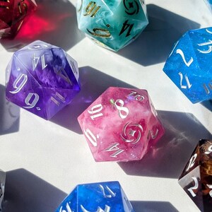 May include: A collection of polyhedral dice in various colors, including blue, pink, purple, and teal. The dice have white or gold numbering and are made of a translucent material, with some containing decorative elements.