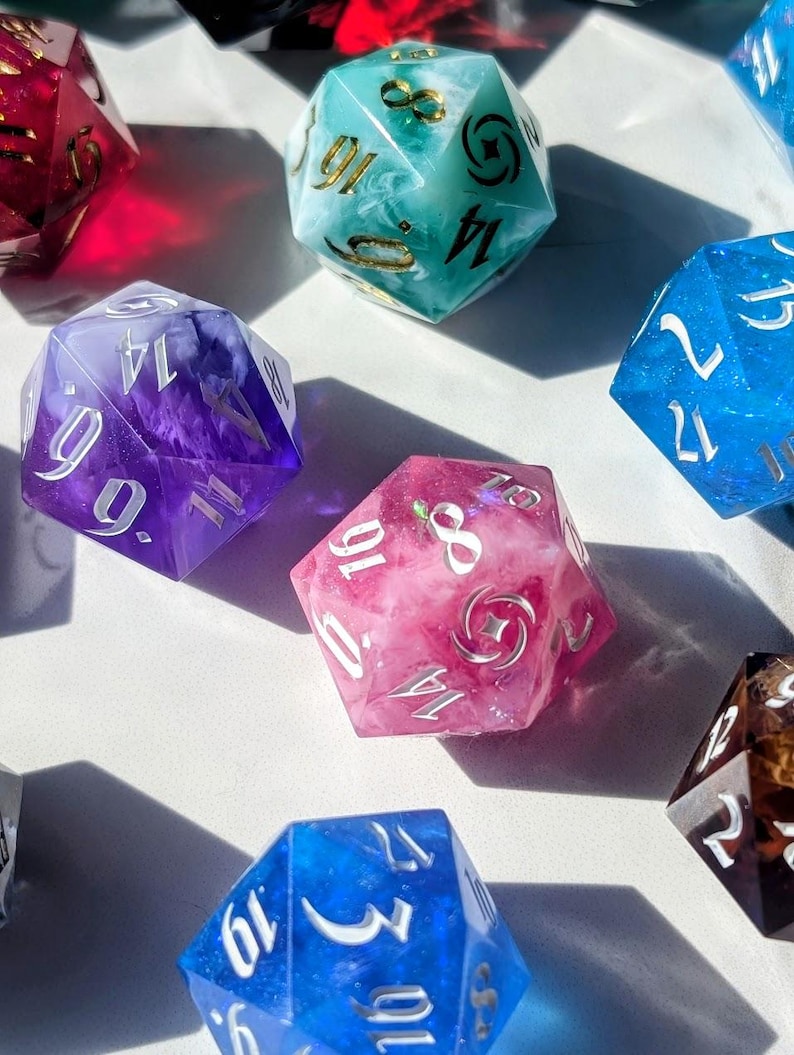 May include: A collection of polyhedral dice in various colors, including blue, pink, purple, and teal. The dice have white or gold numbering and are made of a translucent material, with some containing decorative elements.