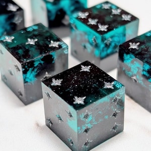 May include: Several resin dice with a dark gray and teal color scheme. Each die features embedded silver star-shaped accents. The dice have a marbled effect, with the teal color swirling within the resin, creating a unique appearance.