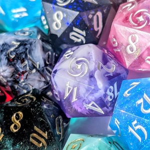 May include: A collection of colorful polyhedral dice in various shades of blue, pink, purple, green, and black. The dice have metallic numbers and are designed for tabletop gaming. The dice are made of resin and have a marbled or glittery appearance.