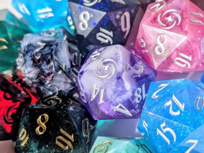 May include: A collection of colorful polyhedral dice in various shades of blue, pink, purple, green, and black. The dice have metallic numbers and are designed for tabletop gaming. The dice are made of resin and have a marbled or glittery appearance.