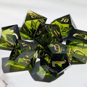 Op de afbeelding: Een set van zeven polyhedrale dobbelstenen met transparante groene hars en goudkleurige cijfers. De dobbelstenen zijn ontworpen voor tabletop-rollenspellen, met elk een verschillend aantal zijden.