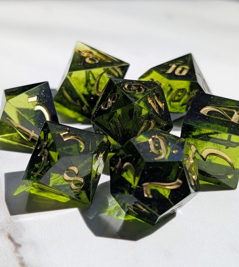 Op de afbeelding: Een set van zeven polyhedrale dobbelstenen met transparante groene hars en goudkleurige cijfers. De dobbelstenen zijn ontworpen voor tabletop-rollenspellen, met elk een verschillend aantal zijden.