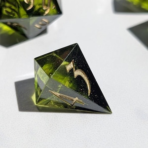 Op de afbeelding: Close-up van een set polyhedrale dobbelstenen. De dobbelstenen zijn doorschijnend groen met goudkleurige cijfers en symbolen. De dobbelstenen zijn gemaakt van hars en zijn ontworpen voor tabletop gaming.
