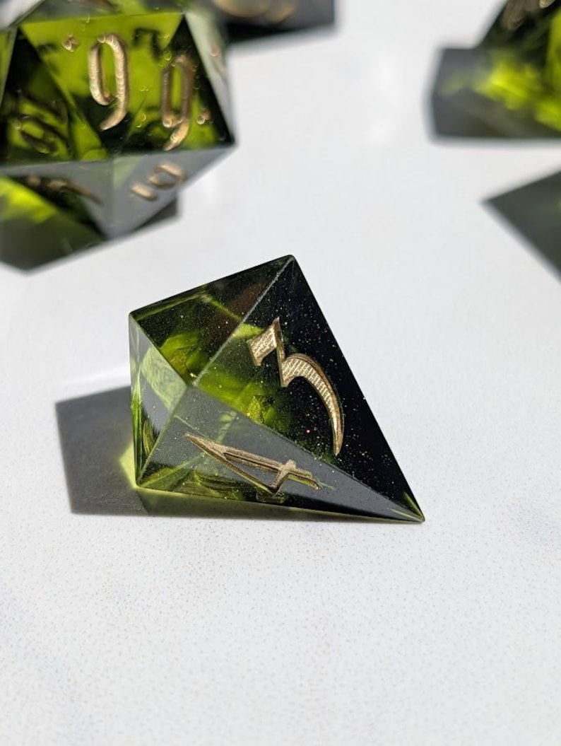 Op de afbeelding: Close-up van een set polyhedrale dobbelstenen. De dobbelstenen zijn doorschijnend groen met goudkleurige cijfers en symbolen. De dobbelstenen zijn gemaakt van hars en zijn ontworpen voor tabletop gaming.