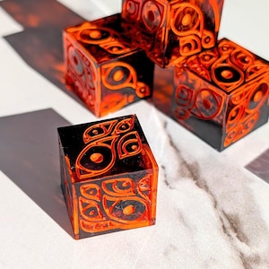 May include: A set of four translucent resin cubes featuring a deep black and vibrant orange swirling pattern. Each cube displays intricate, carved-style eye motifs embedded within the material, creating a layered, dimensional appearance.