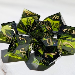 Op de afbeelding: Een set van zeven polyhedrale dobbelstenen met transparante groene hars en goudkleurige cijfers. De dobbelstenen zijn ontworpen voor tabletop-rollenspellen, met verschillende vormen en maten, waaronder een D20, D12, D10, D8, D6 en D4.