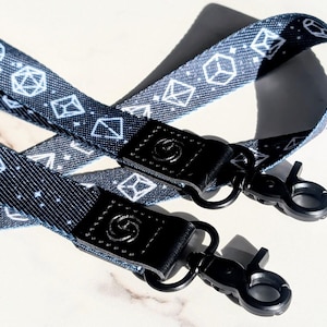 Puede incluir: Dos cordones azul marino con un patrón de dados geométricos blancos. Cada cordón tiene un cierre de cuero negro con un diseño circular y un clip de metal negro. Los cordones están diseñados para sujetar llaves o una tarjeta de identificación.