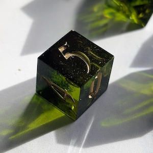 Op de afbeelding: Een handgemaakte, vierkante harsdobbelsteen met het nummer 5 in goud. De dobbelsteen is een mix van groen en zwart met gouden spikkels en ingebedde botanische elementen. De dobbelsteen ligt op een witte ondergrond.