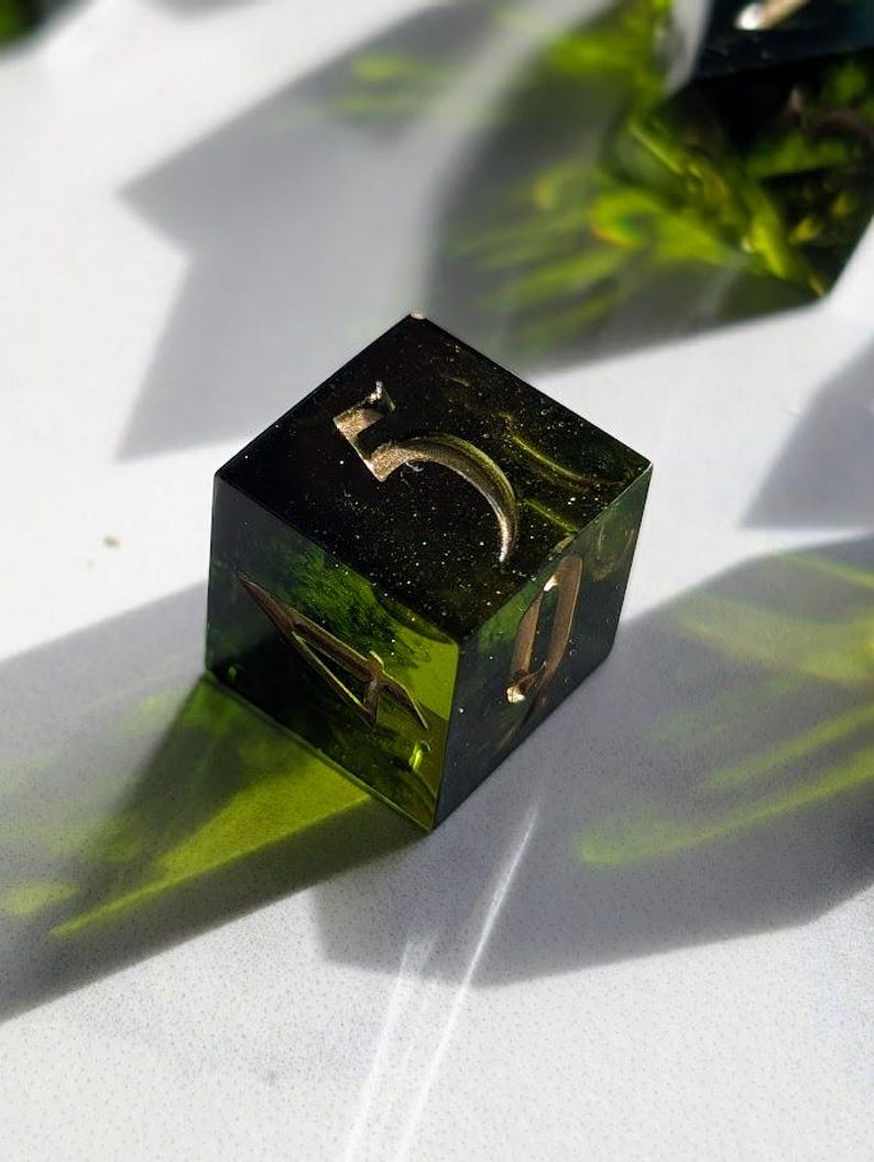 Op de afbeelding: Een handgemaakte, vierkante harsdobbelsteen met het nummer 5 in goud. De dobbelsteen is een mix van groen en zwart met gouden spikkels en ingebedde botanische elementen. De dobbelsteen ligt op een witte ondergrond.