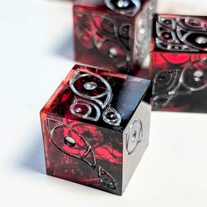以下が含まれることがあります： 銀色のデザインが施された赤と黒のサイコロのクローズアップ。サイコロは正方形で、各面に複雑な模様があります。サイコロは半透明の素材でできており、内部には渦巻く赤い模様が見えます。