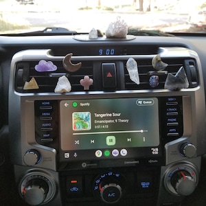 May include: A car's dashboard with a digital clock displaying 9:00. The air vents are decorated with various crystals and other small objects. The car's infotainment system is displaying the song "Tangerine Sour" by Emancipator, 9 Theory, playing on Spotify.