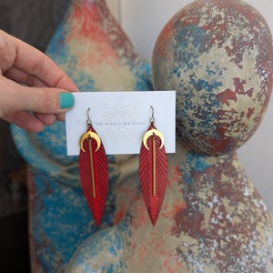 以下が含まれることがあります： 赤い革の羽根のイヤリングに、金の三日月形のアクセントが施されています。イヤリングは、「The Moon & Her Mystic」という文字が書かれた白いカードに飾られています。