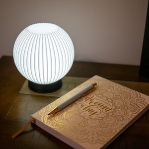 May include: A white, spherical lamp with vertical grooves sits on a dark base. A notebook with the words "dream big" and a pen are next to the lamp. The notebook has a gold floral design.