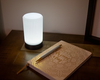 May include: A white cylindrical lamp with vertical lines sits on a wooden table next to a closed notebook with gold lettering that reads "dream big" and a pen.