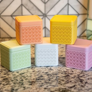 May include: Five colorful square boxes with a geometric pattern on the sides. The boxes are in shades of orange, yellow, pink, green, and white. The lids are solid colors. The boxes are on a speckled surface.